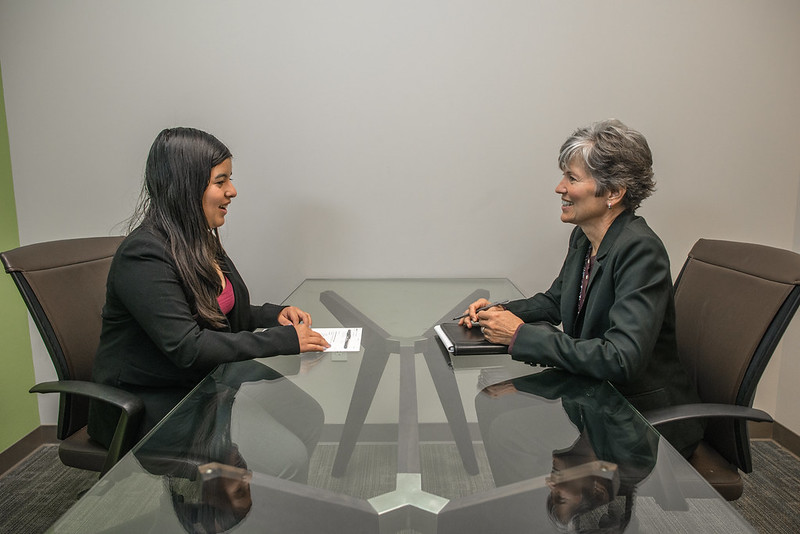 Duas mulheres, uma de frente para a outra, e sentadas, diante de uma entrevista de emprego